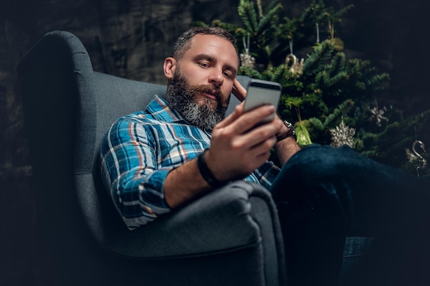 Maschio barbuto di mezza età che utilizza smartphone su sfondo natalizio decorato.