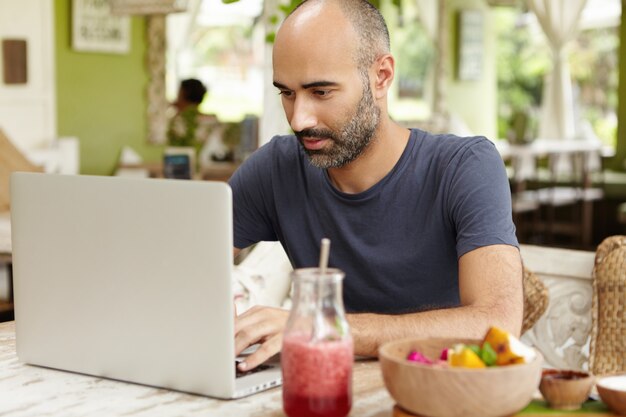 Bearded middle-aged self-employed man sitting at cafe in front of generic laptop and looking at screen with serious and concentrated expression while working remotely on his project, using free wi-fi