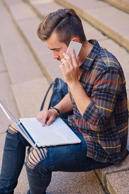 ひげを生やした現代の男性が階段に座り、スマートフォンとラップトップを使用して話します。