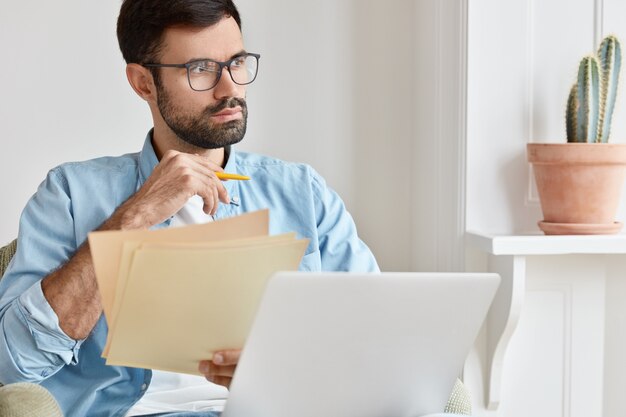 Bearded pensive man works from home, counts financial data, holds paper documents