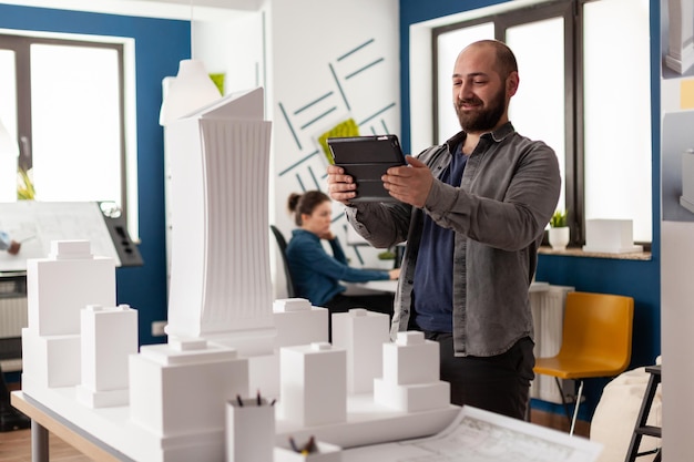 수염을 기른 전문 건축가는 건축 사무소의 흰색 거품 건물 모델 앞에서 태블릿을 보고 있습니다. 디지털 장치의 청사진을 마천루의 규모 설계와 비교하는 엔지니어.