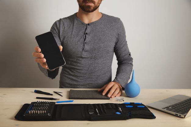 Bearded professional shows repaired fixed smartphone after service replacement, above his specific tools in toolkit bag near laptop on wooden white table