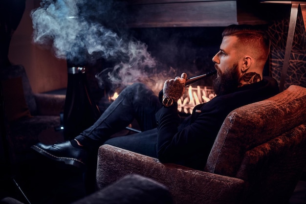 Bearded relaxed man is sitting on the armchair and smoking hookah near fireplace.