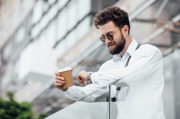 A bearded serious stylish manager looking to his watch on the streets of the city near modern office centre Man drinks coffee An employee looks at the time