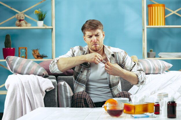 Bearded sick man with flue sitting on sofa at home and measuring body temperature. The winter, illness, influenza, pain concept. Relaxation at Home. Healthcare Concepts.