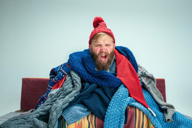 Bearded sick man with flue sitting on sofa at home or studio covered with knitted warm clothes. Illness, influenza concept. Relaxation at Home. Healthcare Concepts.