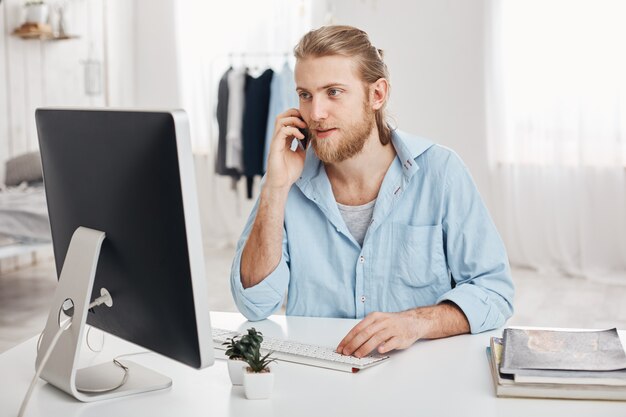 Bearded skilled young fair-haired businessman works on new project, sits in front of screen, has phone conversation, discusses financial report with businesspartner. Office worker chats with boss