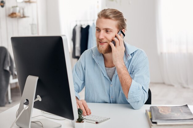 Bearded skilled young fair-haired copywriter works on new article, types on keyboard, has phone conversation, discusses new project with businesspartner. Successful businessman has important call.