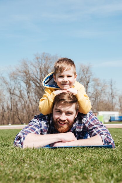 Bearded smiling father lies with little son in the park.