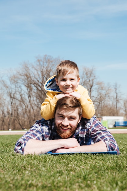 Bearded smiling father lies with little son in the park.