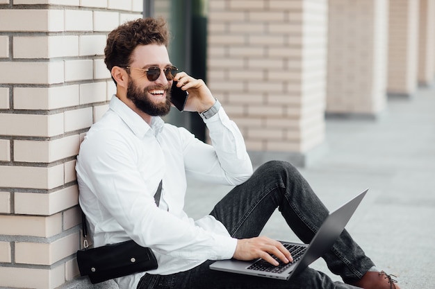 A bearded, smiling, stylish man sitting on the flour on the streets of the city near modern office centre and workin with his laptop and calling phone