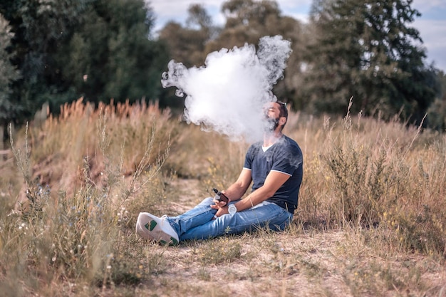 Bearded smoker blowing a lot of smoke using vape electronic cigarette in nature. electronic cigarette. Premium Photo