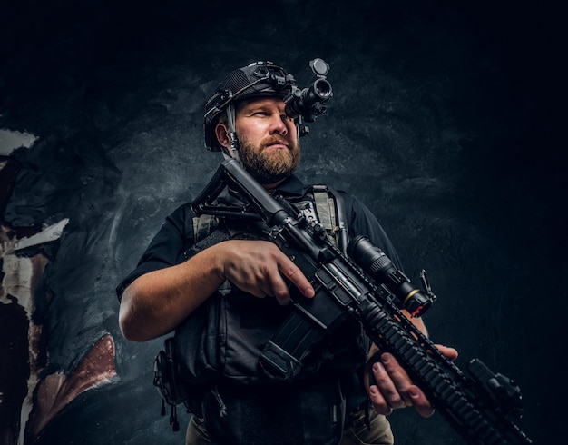 Bearded special forces soldier or private military contractor holding an assault rifle and observes the surroundings in night vision goggles. Studio photo against a dark textured wall