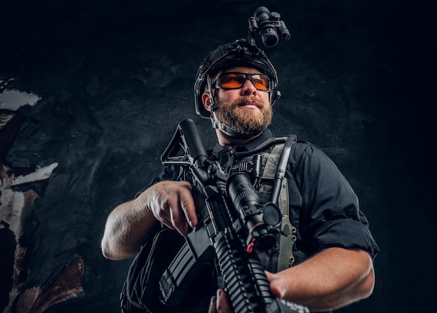 Bearded special forces soldier wearing body armor and helmet with night vision holding an assault rifle. Studio photo against a dark textured wall