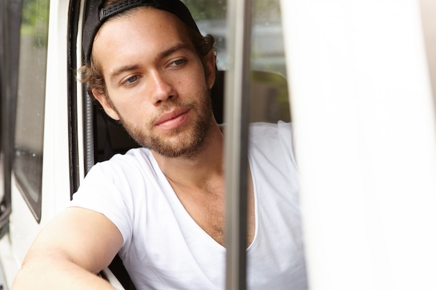 Free photo bearded student in snapback driving white jeep, sticking his head and elbow out of open window, looking ahead at road while stopping at crossroads for red light