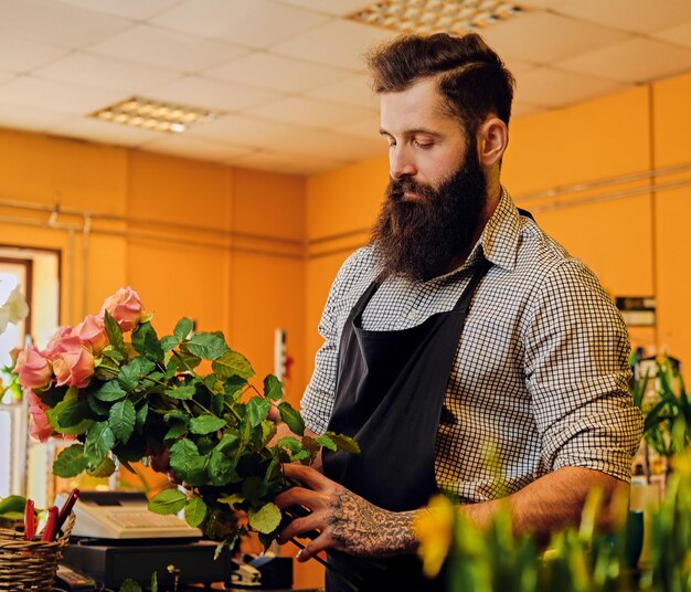 The bearded stylish flower seller holds pink roses in a market shop.