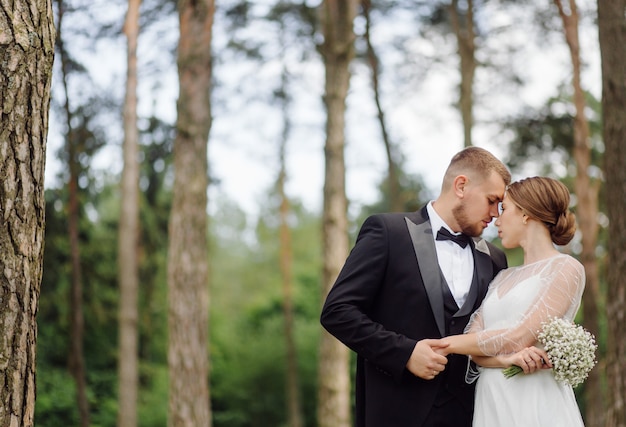 スーツを着たひげを生やしたスタイリッシュな新郎と、花束を手にした白いドレスを着た美しい金髪の花嫁が、松林の中で立って抱きしめています。