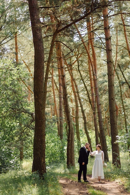 A bearded, stylish groom in a suit and a beautiful blonde bride in a white dress with a bouquet in her hands are standing and hugging in nature in the pine forest.