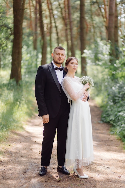 A bearded, stylish groom in a suit and a beautiful blonde bride in a white dress with a bouquet in her hands are standing and hugging in nature in the pine forest.