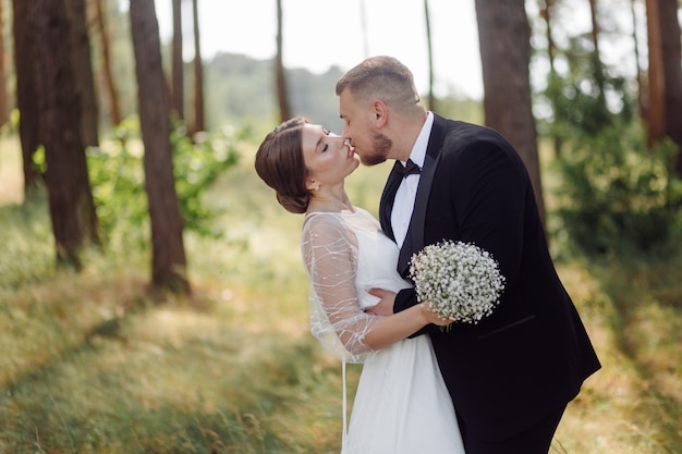 Uno sposo barbuto ed elegante in abito e una bella sposa bionda in abito bianco con un bouquet in mano sono in piedi e si abbracciano nella natura nella foresta di pini.