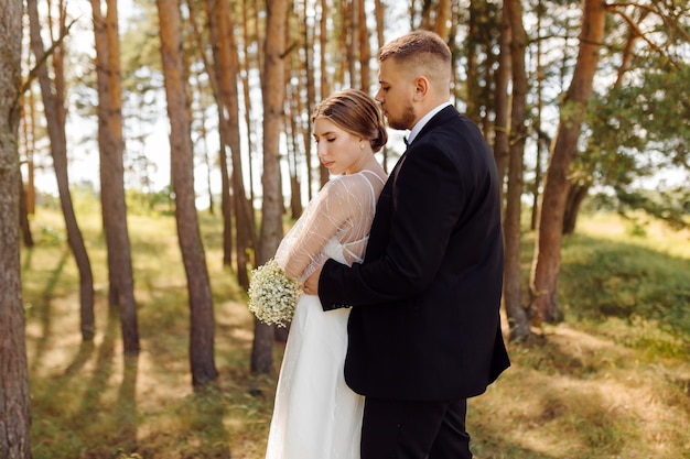 A bearded, stylish groom in a suit and a beautiful blonde bride in a white dress with a bouquet in her hands are standing and hugging in nature in the pine forest.