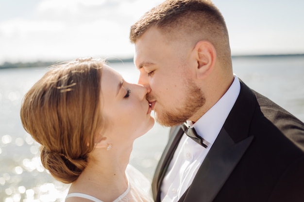 A bearded, stylish groom in a suit and a beautiful blonde bride in a white dress with a bouquet in her hands are standing and hugging in nature in the pine forest.