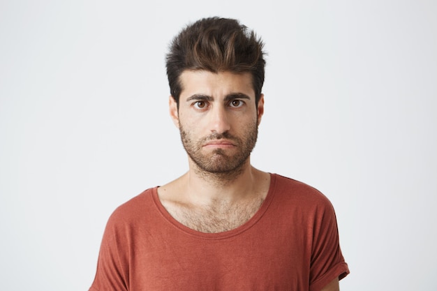 Bearded stylish male having displeased look standing in casual cloth against white wall. Young man having some problems looking with unsatisfaction and discontent 