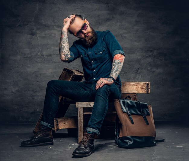 Bearded stylish male in a jeans and sunglasses sits on wooden box over grey background.
