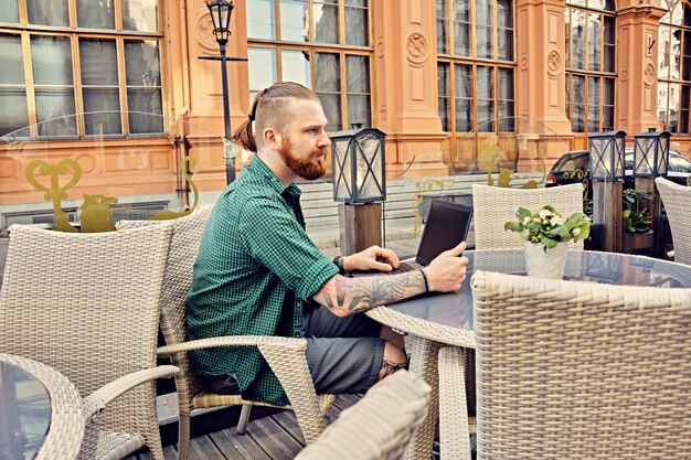 Бородатый татуированный мужчина с ноутбуком в открытом уличном кафе в старом городе.