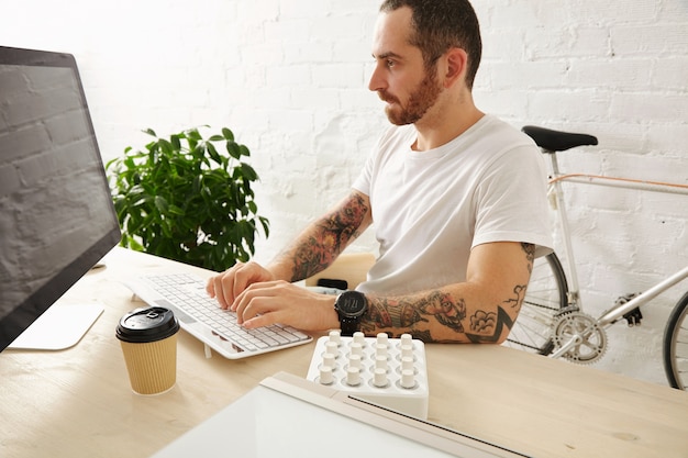 Bearded tattooed man in blank white t-shirt works on his computer at home, side view, summer time