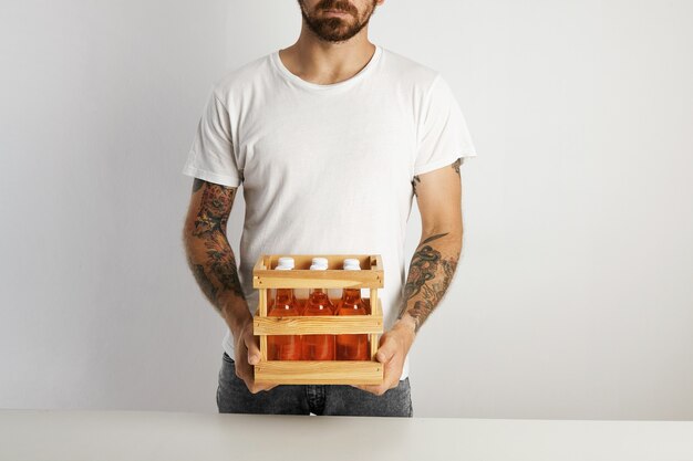 白い壁にクラフトラガービール飲料のマークされていないガラス瓶6本と小さな木枠を保持しているひげを生やした入れ墨の男