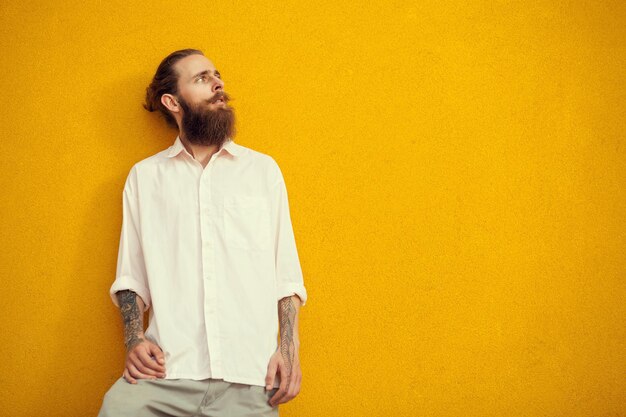 Бородатый и татуированный мужчина позирует на желтой стене снаружи. Мода и стиль