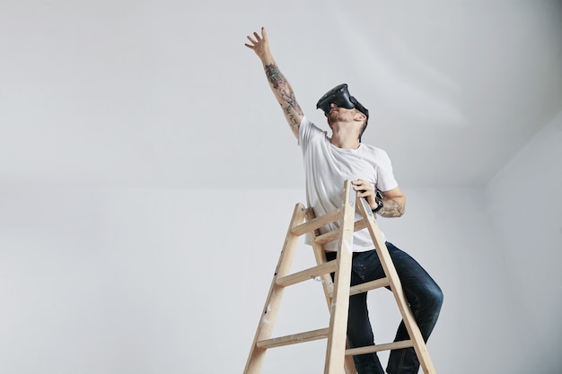 Бородатый и татуированный молодой человек в белой футболке без надписи и очках виртуальной реальности стоит на лестнице и тянется к чему-то