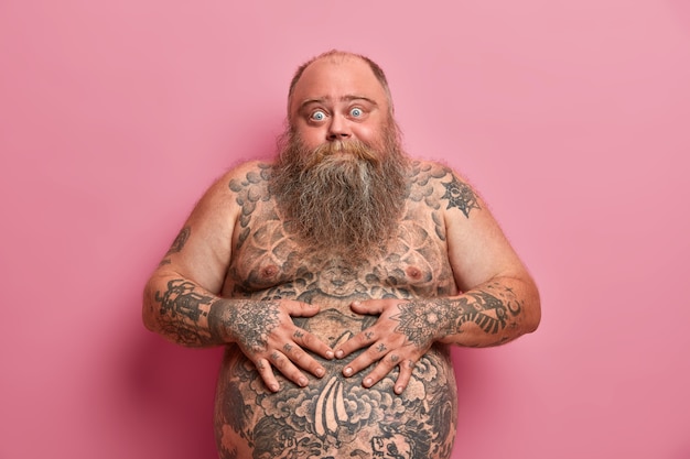 Bearded thick guy keeps hands on big tattoed belly, has bugged eyes, has thick beard, poses against pink wall. Naked overweight adult man with large tummy, asks advice how to loose weight