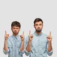 Free photo bearded two men with sullen expressions, point upwards with both index fingers, being discontent