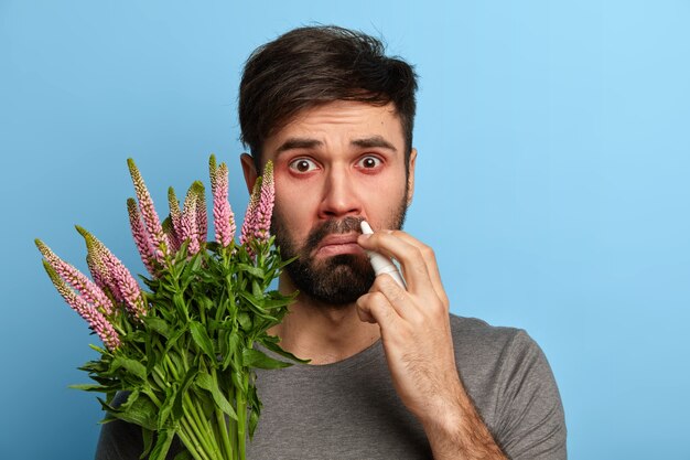 Бородатый нездоровый мужчина страдает сезонной аллергией, брызгает в нос каплями в нос, держит растение, будучи чувствительным к аллергену, позирует у синей стены. Медицинская концепция