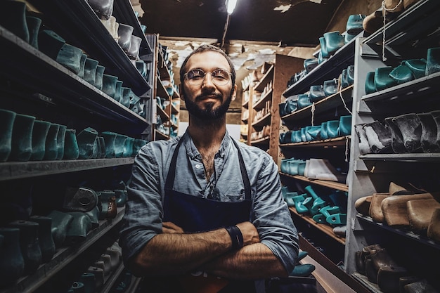 Bearded young cobbler in glasses is posing for photographer at dark shoe form storage.