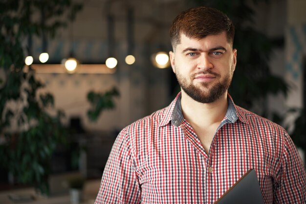 Бородатый молодой человек в повседневной рубашке, стоя с ноутбуком в руках