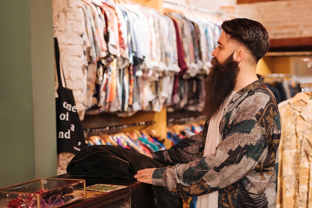 Bearded young man at the counter looking at clothes hanging on rail