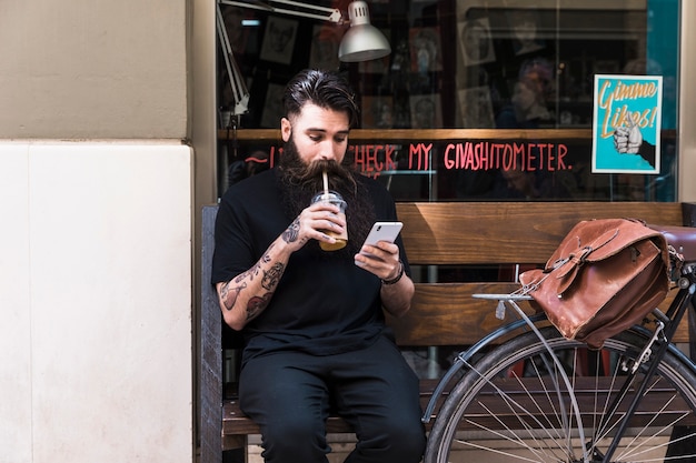 Free photo bearded young man sitting on bench outside the cafe drinking chocolate drink using mobile phone