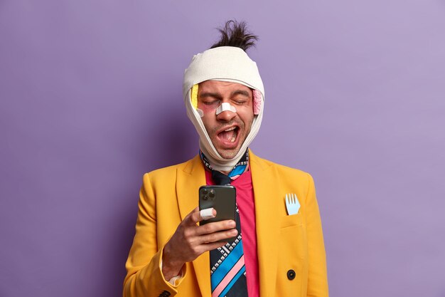 Beaten bruised man feels bored at home during rehabilitation period, uses smartphone and yawns with sleepy expression, injured after serious accident, dressed in bright clothes, poses indoor