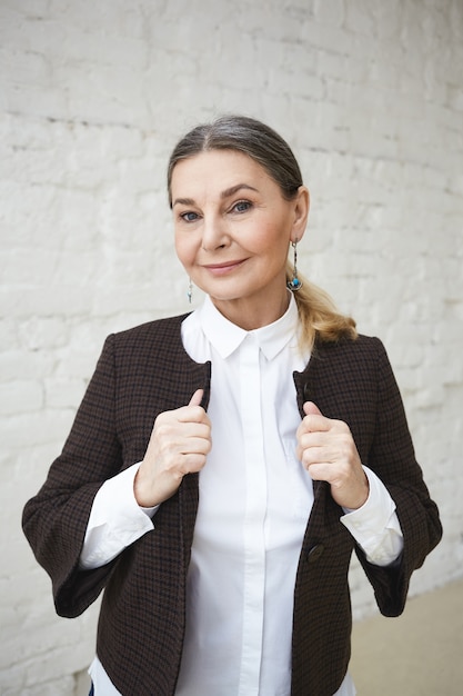 Beaty, style, fashion and age concept. Waist up shot of beautiful gray haired 50 year old female posing indoors, standing at white brick wall, adjusting her stylish outfit, going to have meeting