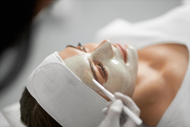 Косметолог, применяя специальную белую маску на лице для женщины