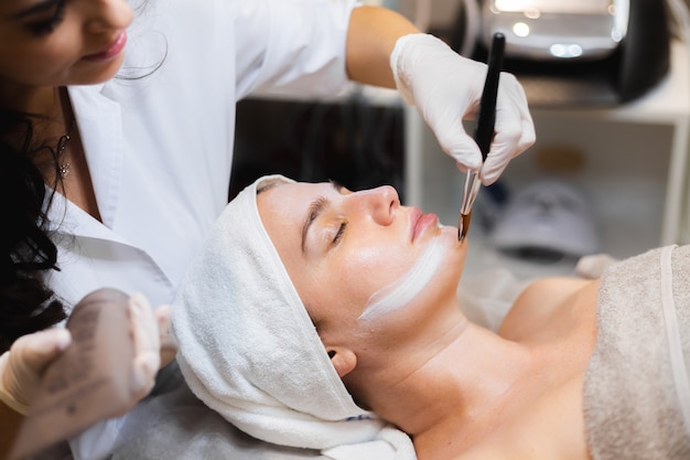 Beautician with a brush applies a white moisturizing mask to the face of a young girl client in a spa beauty salon