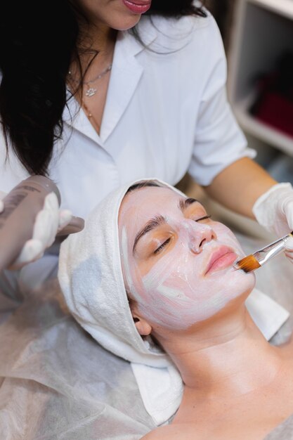 Косметолог с кисточкой наносит белую увлажняющую маску на лицо молодой девушки-клиента в спа-салоне красоты