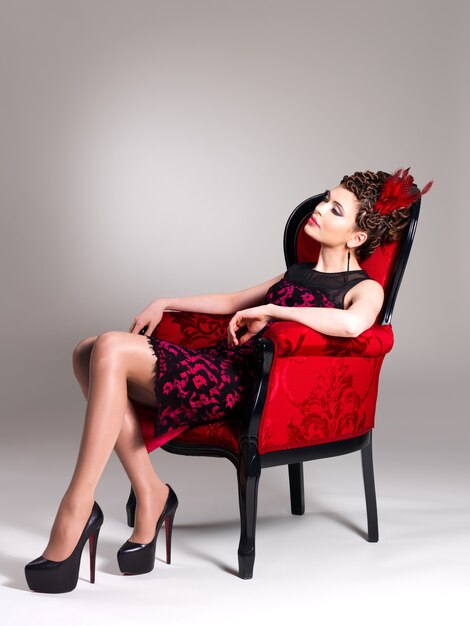 Beautiful adult woman with fashion hairstyle and red armchair poses at studio