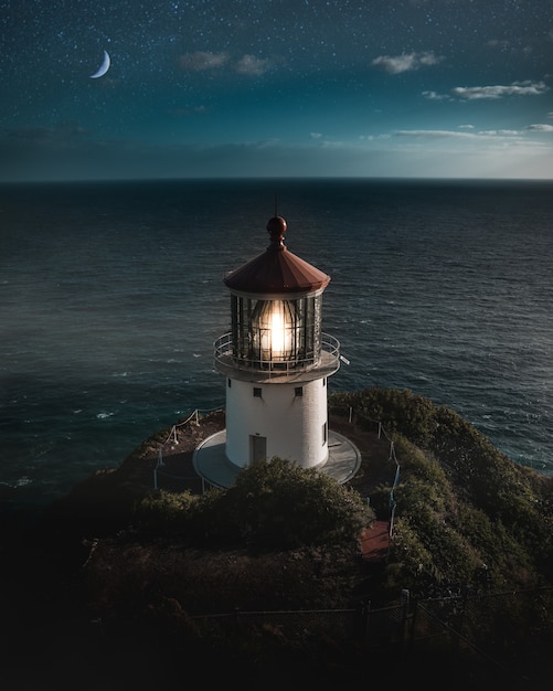 Free photo beautiful aerial shot of a lit lighthouse on a green hill with the half-moon in the night sky