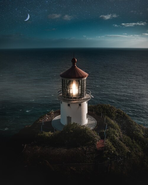 Beautiful aerial shot of a lit lighthouse on a green hill with the half-moon in the night sky