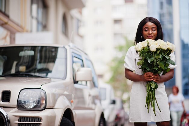 Beautiful african american girl holding bouquet of white roses flowers on dating in the city Black businesswoman with bunch of flowers near car