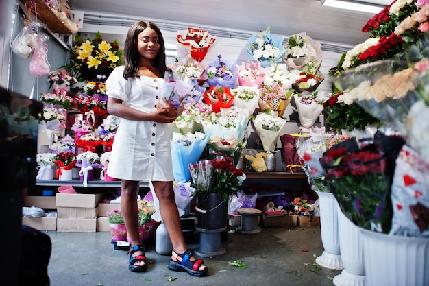 Beautiful african american girl in tender white dress with bouquet flowers in hands standing against floral background in flower shopBlack female florist